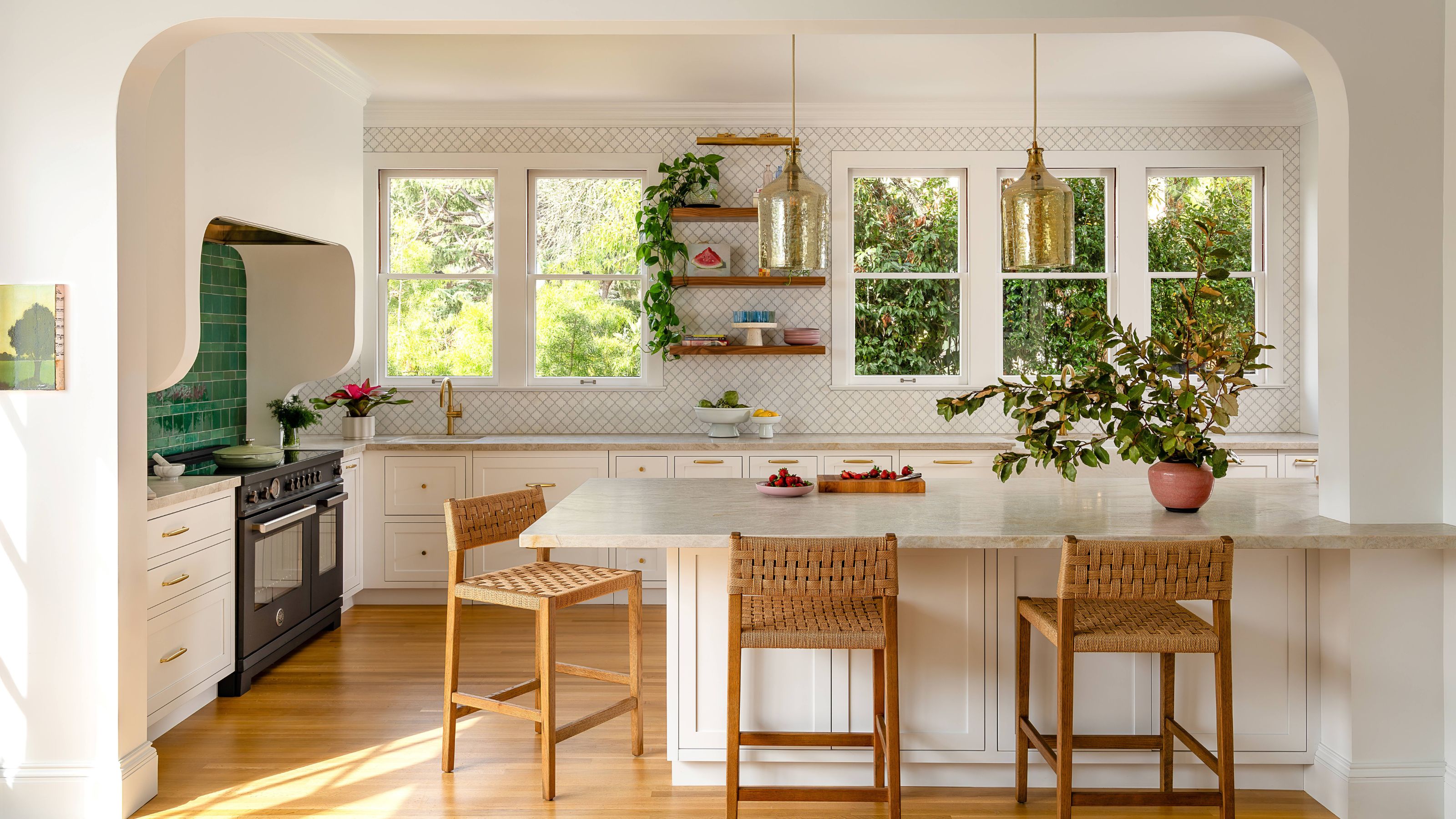 a white kitchen with a large range cooker with green tiles, a wallpapered wall, and a peninsula island with woven wooden bar stools