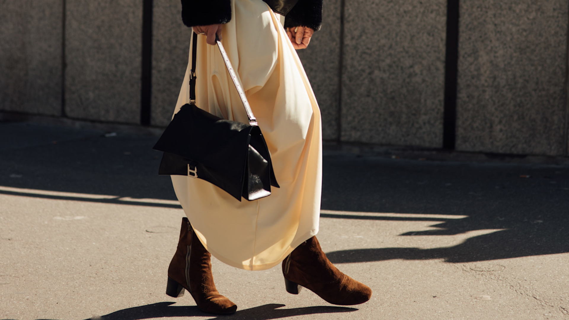 a woman walking on a sidewalk wearing a black fur sleeve jacket, white skirt, brown suede booties, and a black handbag.