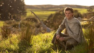Robert Aramayo sits on the grass in costume as Elrond in The Rings of Power.