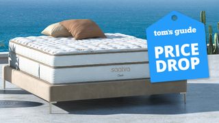 A Firm Saatva Classic Mattress on a bed frame outdoors on a platform by the sea, a Tom's Guide price drop deals graphic (right)