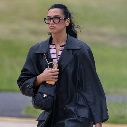 Dua Lipa travels with a leather trench coat and double bags by Chanel and Hermes, plus a pair of Margiela Tabi flats