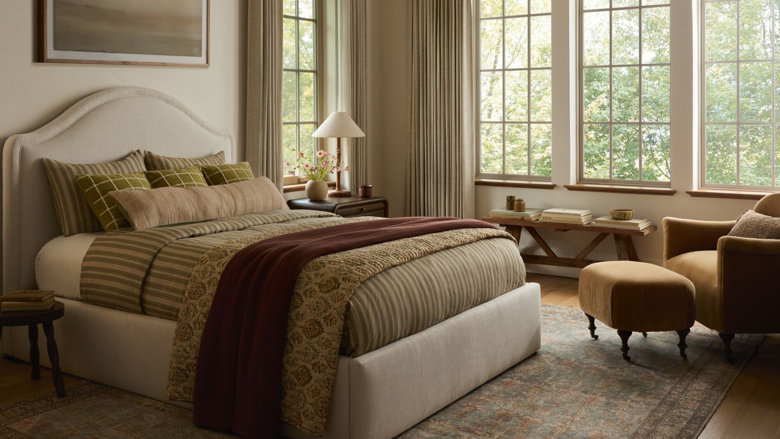 a bedroom with neutral colors, an upholstered bedframe with green beddings, an accent chair with a small ottoman, and a wall of windows with curtains hanging in front