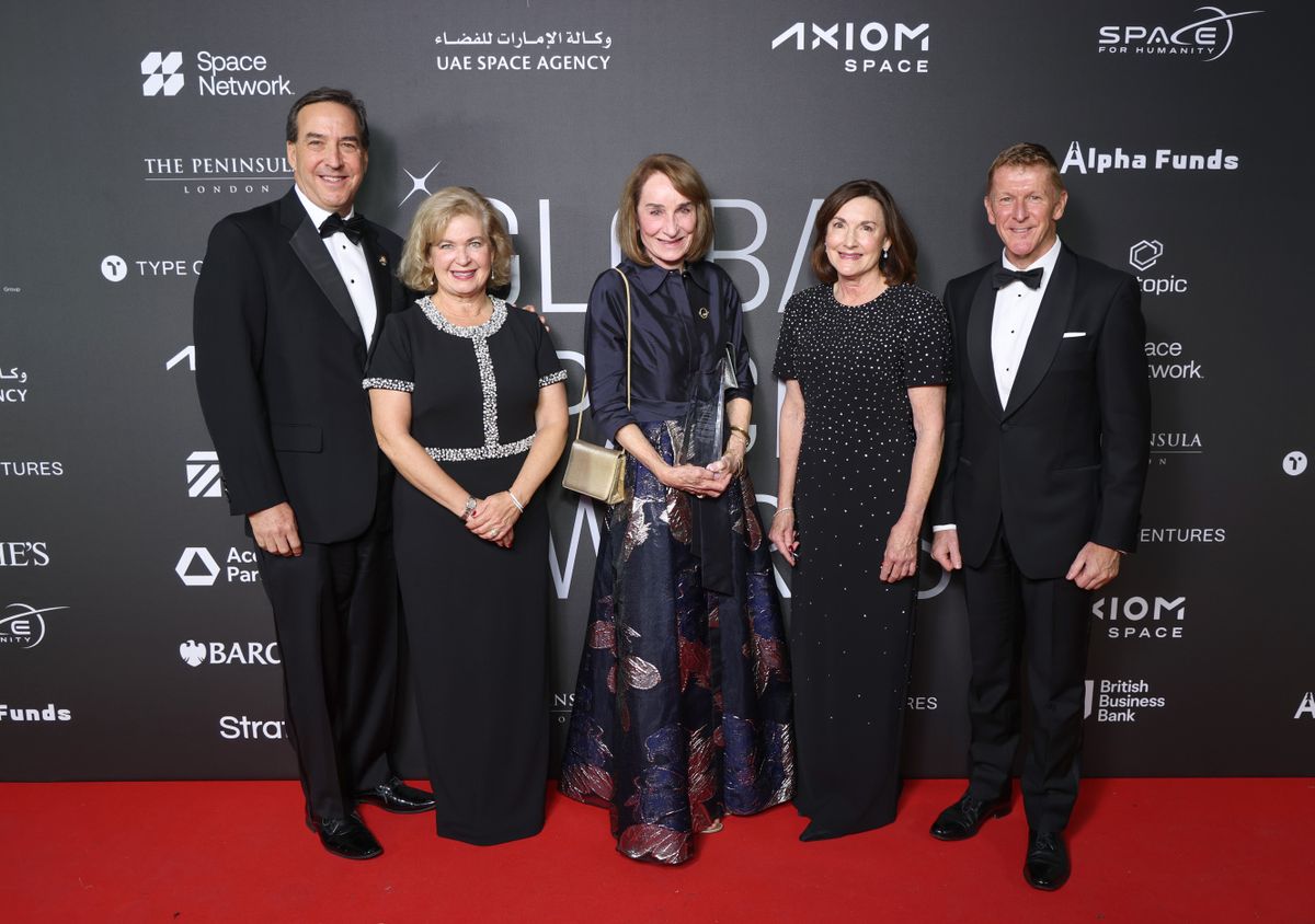 Un grupo de cinco personas con esmoquin y vestidos formales se encuentran sobre una alfombra roja.
