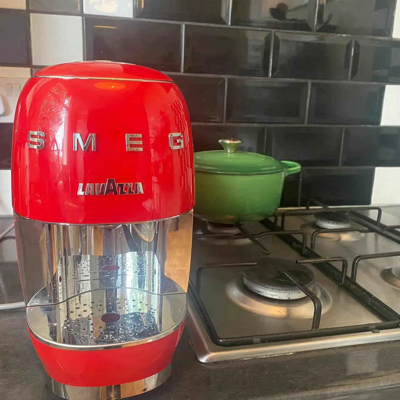 A red SMEG pod coffee machine on a worktop with a green pan on the hob beside/behind