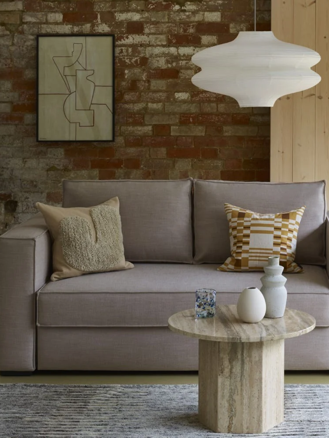 A beige sofa bed from Heal's in a living room with a stacked pendant, a stone coffee table, vases, and a framed print