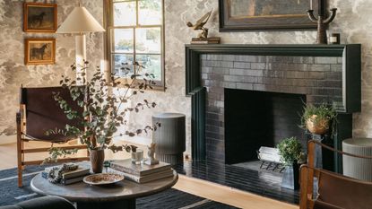 Image of a living room with fine line black and white wallpaper that has cloud motifs. There is a large black tile fireplace with a round coffee table in front of it, a black rug, and two leather and wood accent chairs, and a white floor lamp.