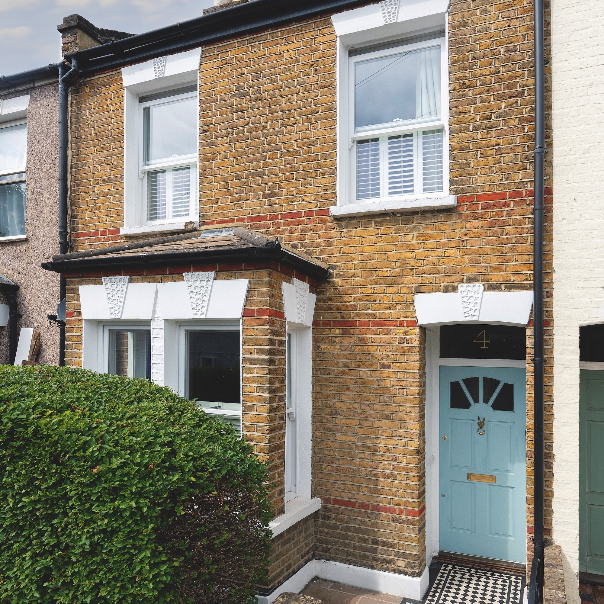 exterior of brick victorian mid terrace house in London with pale blue front door, black and white tiled path, and hedge