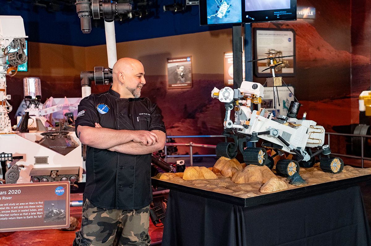 a man in a black polo shirt stands in front of a life-size model of nasa&#039;s perseverance mars rover