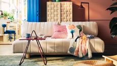 A cream sofa bed in an orange living room with two colorful cushions and a throw with a red side table in front of it with a water jug and cup and a woven wooden cabinet behind it next to a blue door