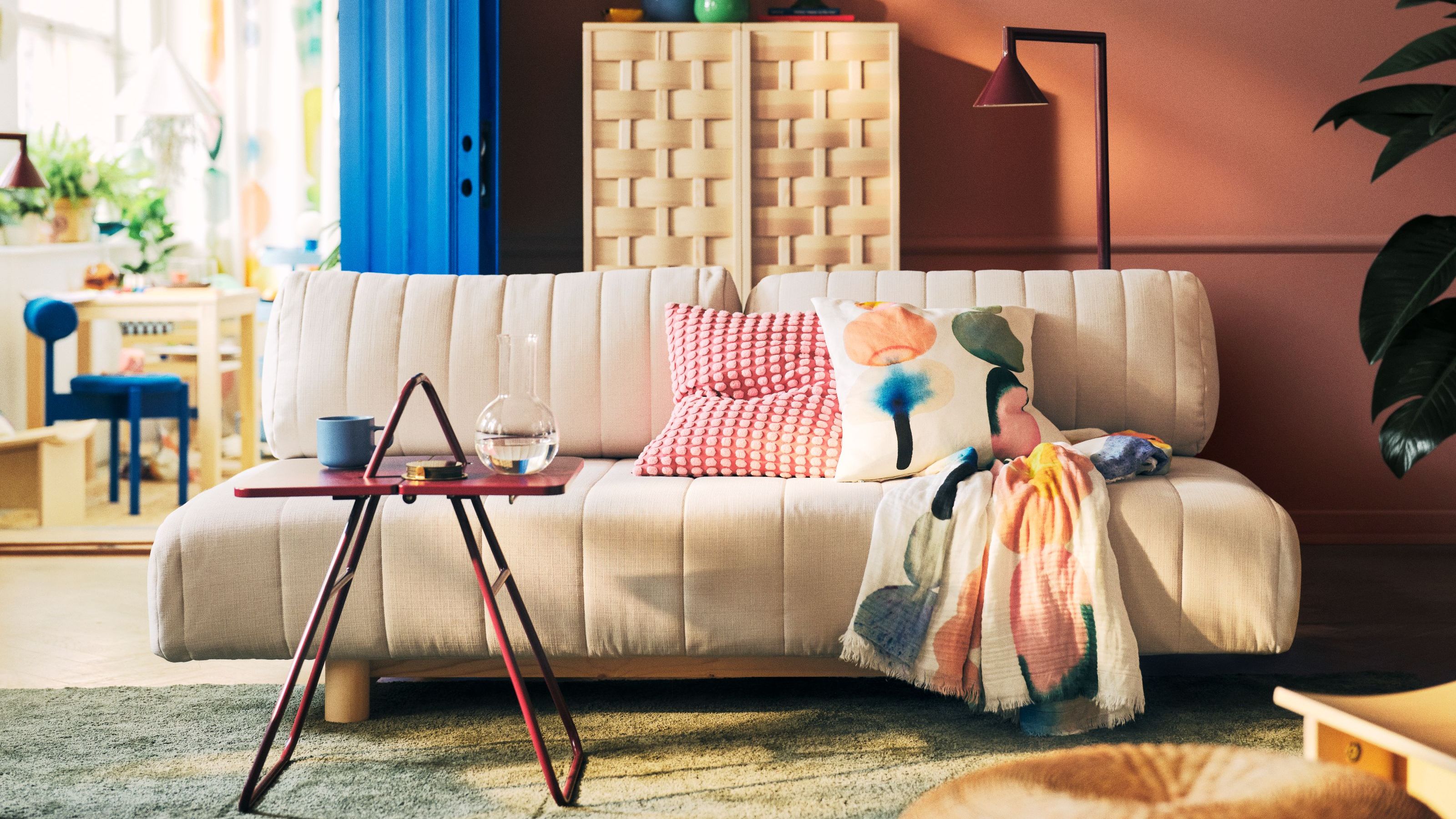 A cream sofa bed in an orange living room with two colorful cushions and a throw with a red side table in front of it with a water jug and cup and a woven wooden cabinet behind it next to a blue door