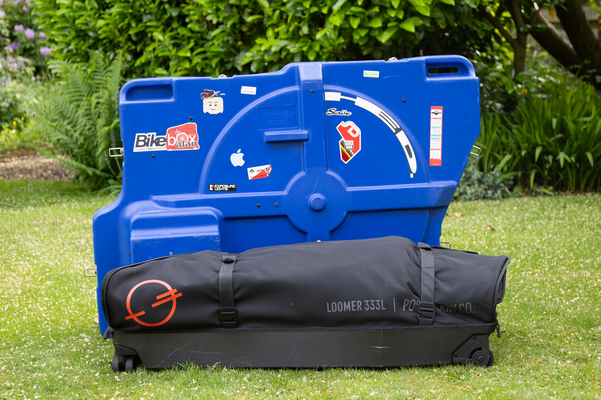 black bike box packed down standing on a lawn in front of a blue hard case bike box