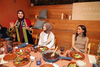 Meghan Markle sitting at a dinner table covered in plates and food watching a woman standing up and talking who is wearing traditional Afghan dress