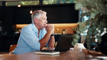 Portrait of thoughtful mature man with laptop sitting at wooden table 