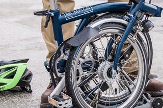 This image is of a folding bike, folded up. There is a green helmet on the ground next to the bike a behind the bike you can see a pair of legs in brown trousers with brown boots on.