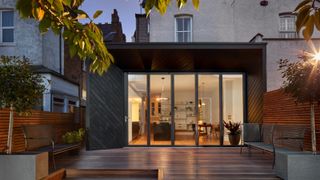 evening shot of wrap around extension with bi fold doors, black cladding, timber deck and white rendered house in background