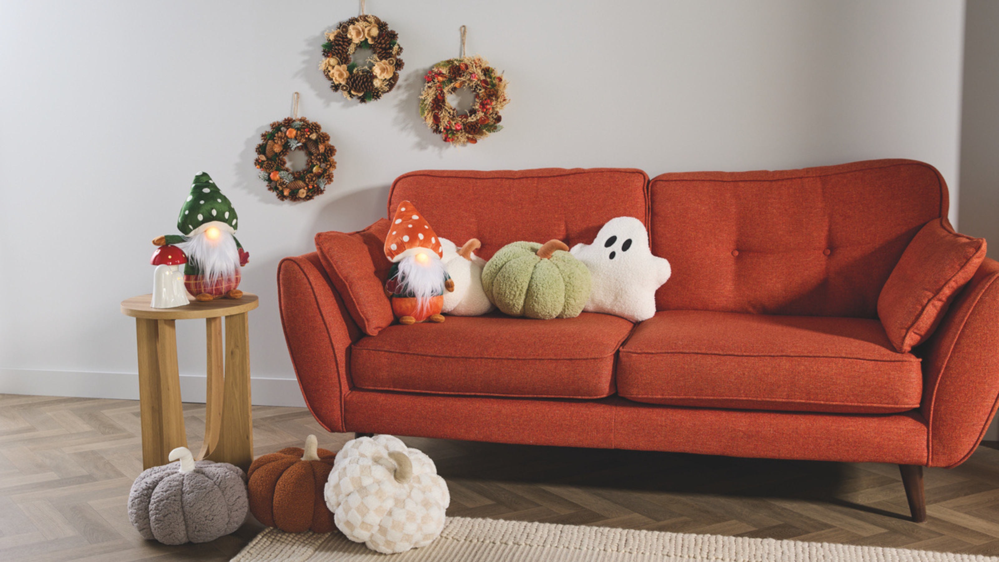 Aldi autumnal decor on a red sofa. There are plush pumpkins, ceramic lights, a ghost pillow, gons and wreathes.