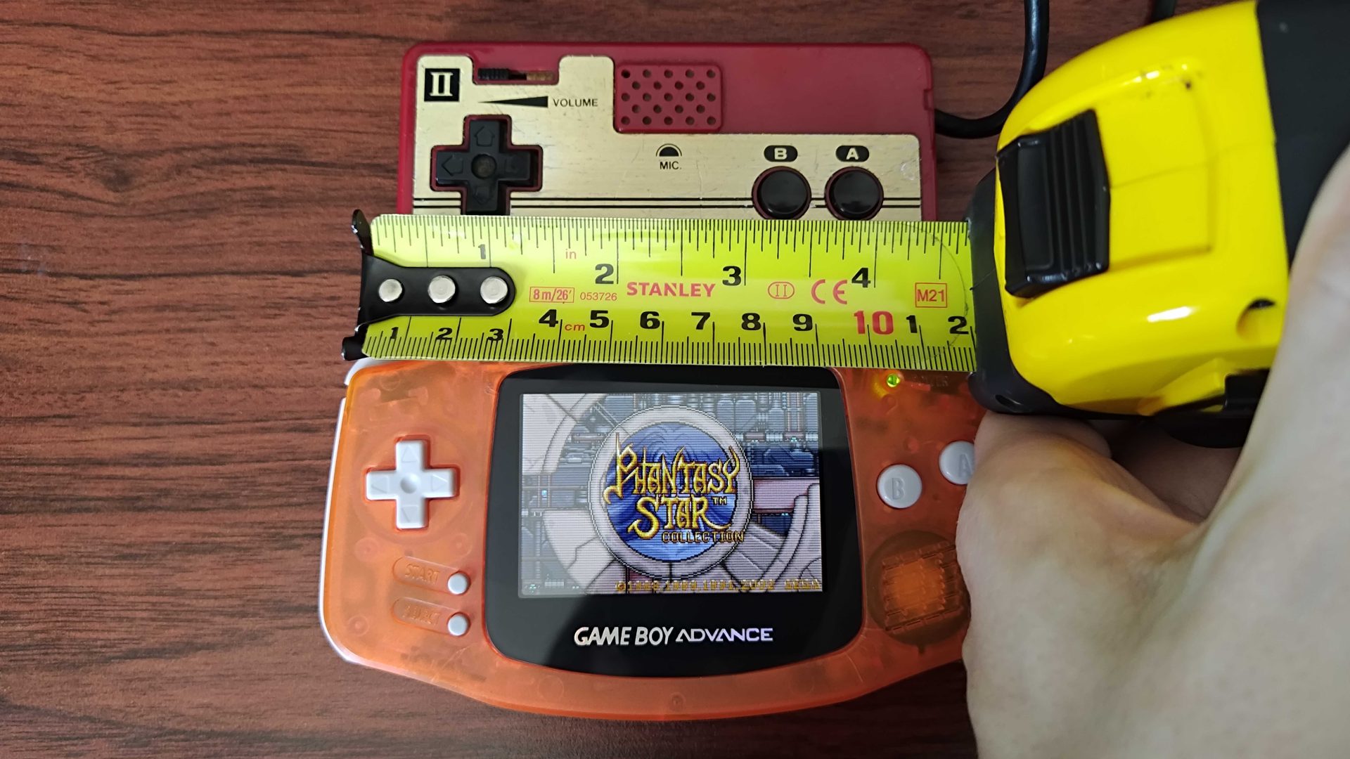 Hand holding tape measure between orange GBA and a detached Famicom controller.