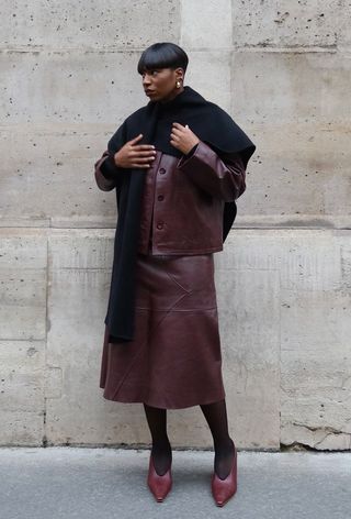 A French woman standing outside wearing a black cape scarf over a red leather jacket with a red leather skirt, black tights, and red pumps.