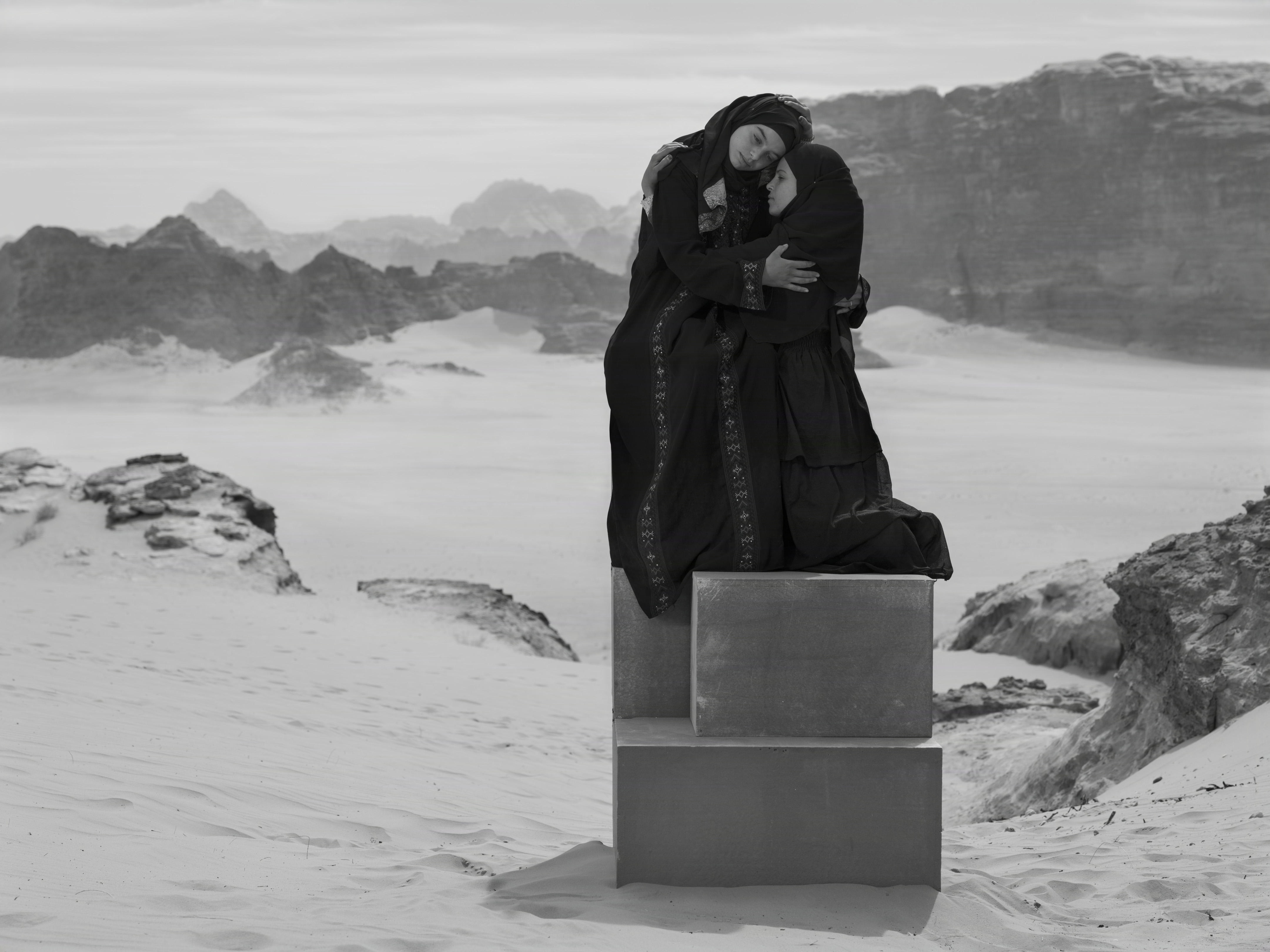 Two women in dark, traditional clothing embrace while seated atop stacked stone blocks in a wide desert valley.