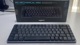 Det trådlösa tangentbordet Logitech MX Mechanical Mini står på ett vitt bord framför sin tillhörande förpackning.