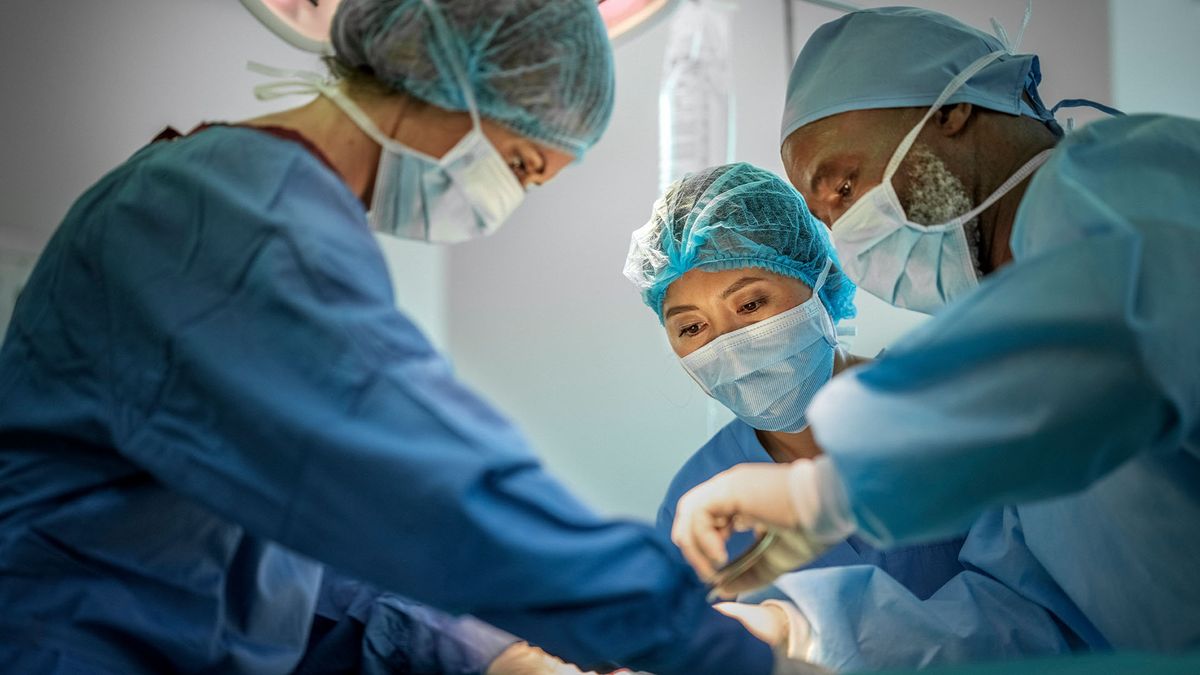 three doctors wearing blue surgical scrubs, face masks and hair nets pictured bent over an operating table, which is not shown