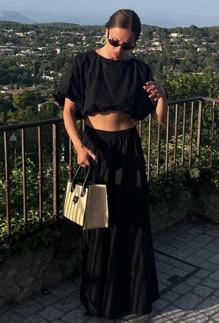a black maxi skirt outfit showcased on a woman wearing sunglasses with green resin hoops, a cropped black crop top, a black tiered maxi skirt and a raffia handbag