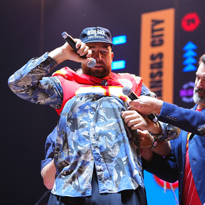 Travis Kelce puts on a jersey as he stands onstage with Rob Riggle and Paul Rudd during 2024 Big Slick Celebrity Weekend on June 01, 2024 in Kansas City, Missouri.