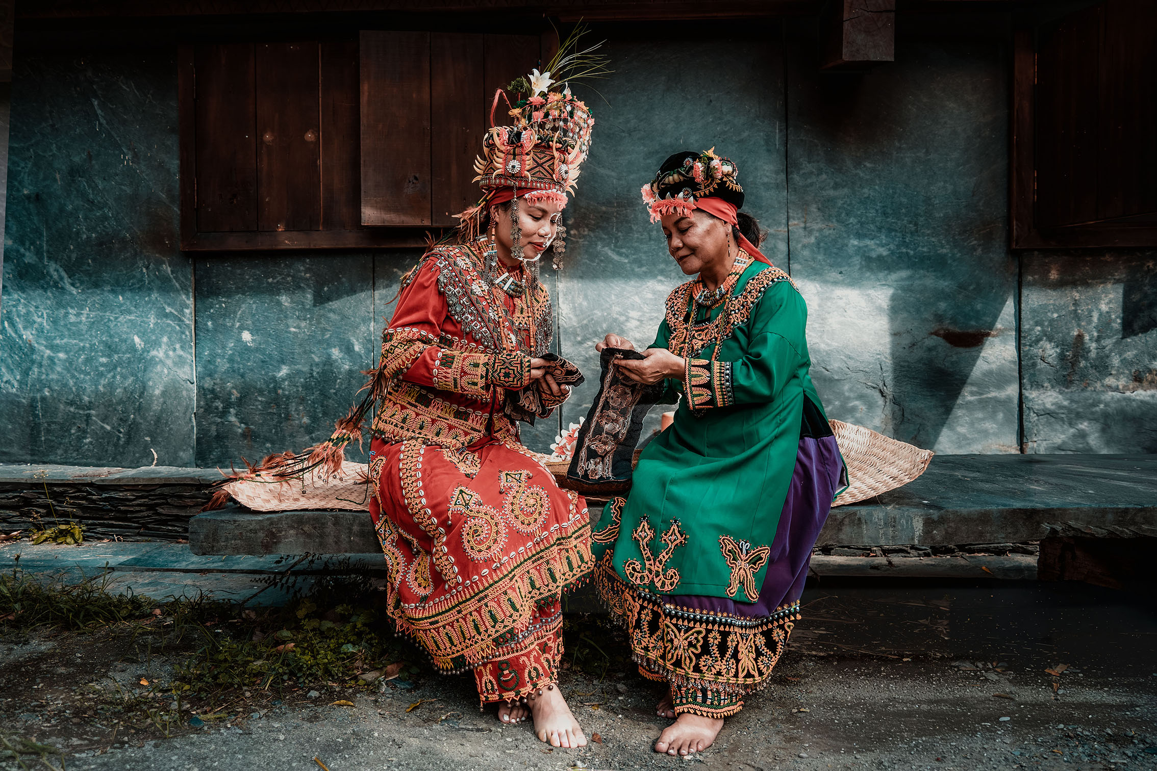 Two people dressed in colorful traditional attire and headdresses sit barefoot on a stone ledge, holding fabric pieces