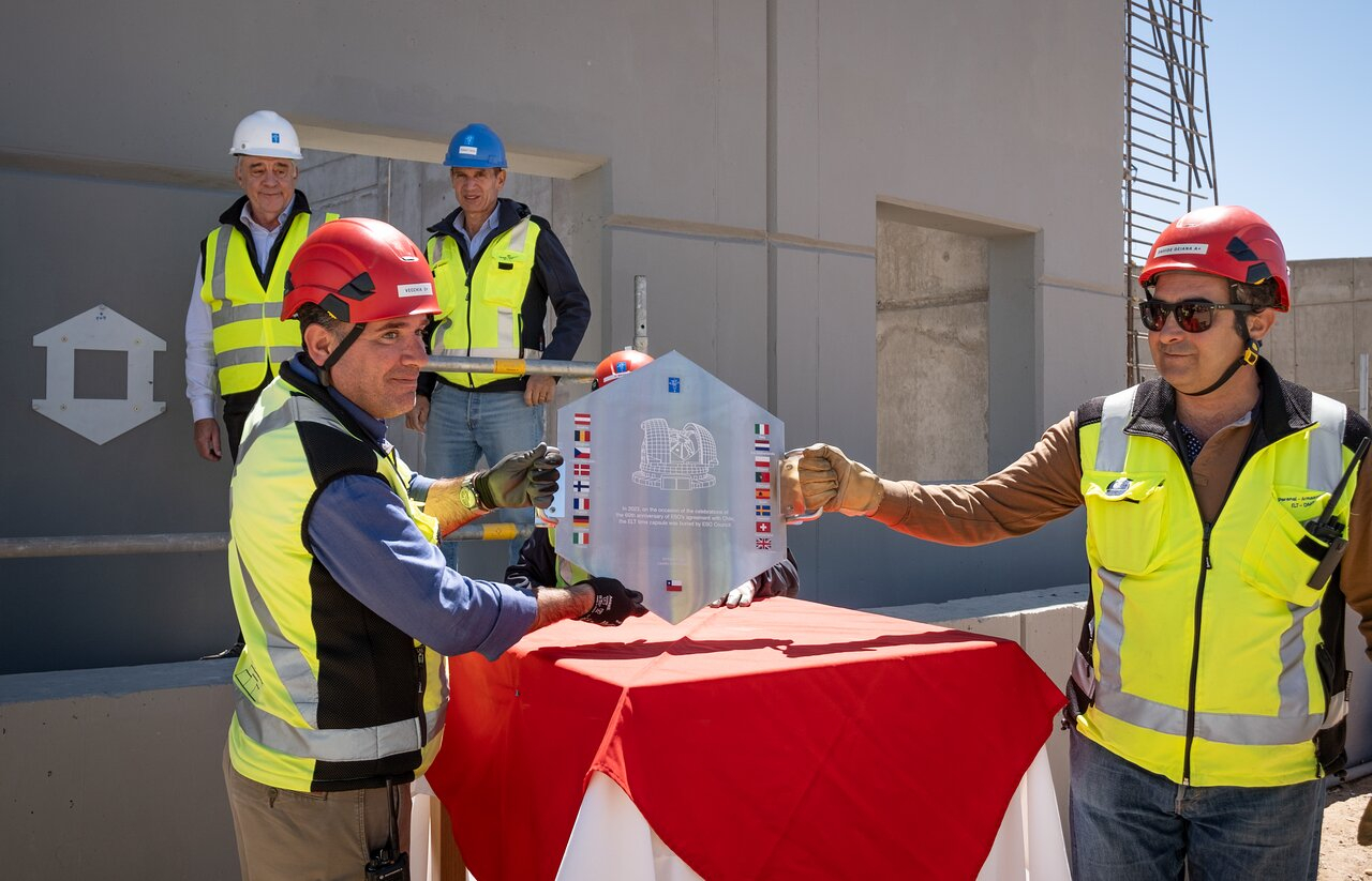 Two figures wearing high-visibility jackets and helmets stand flanked by silver hexagonal shields. Behind him, three people also wearing construction equipment are watching.