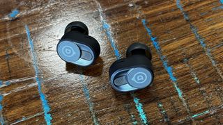 The Devialet Gemini II wireless earbuds shown on a textured wooden background
