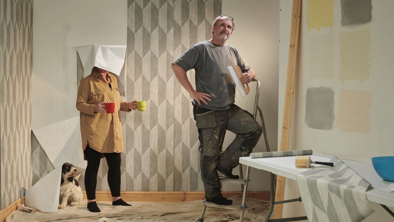 A woman has falling wallpaper draped over her head while she stands behind a man who looks very pleased with the work he&#039;s done.