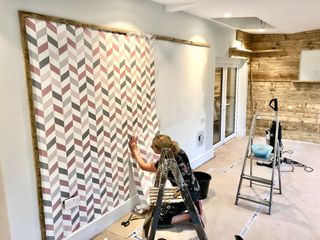 A woman wallpapering a wall with chevron pink and grey chevron patterned paper
