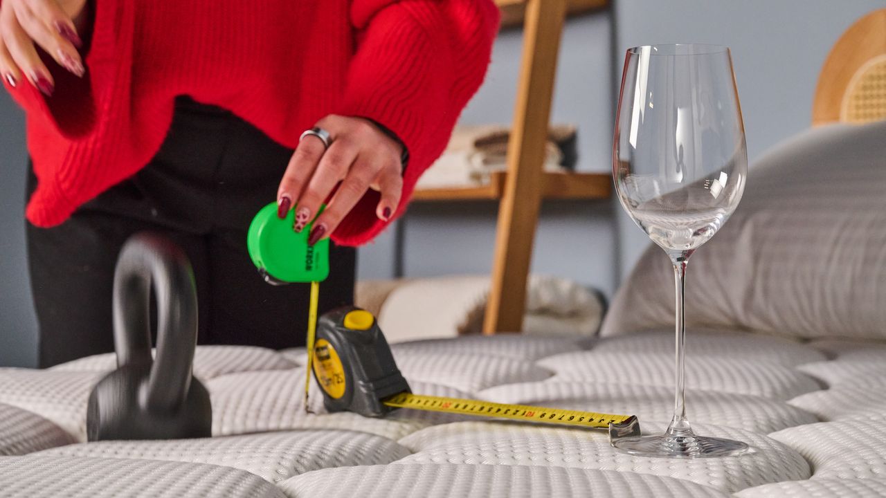 One of the Tom's Guide mattress testers holds a green and yellow measuring tape near a heavy black kettlebell and a wine glass during a motion isolation weight drop test