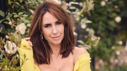 Alex Jones pictured outdoors in front of some flowers, wearing a yellow cardigan with fresh, summery makeup 