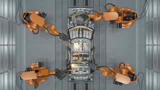 A top-down shot of a car being made using smart manufacturing robot arms, against a grey metal floor. There are four of the robot arms and they are orange.