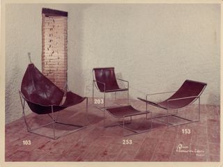 An archive photograph showing three lounge chairs and one ottoman displayed in a circle inside a minimally-decorated home, designed by Odile Mer and originally produced by Prisunic. The furniture features chrome structures and cognac leather seats