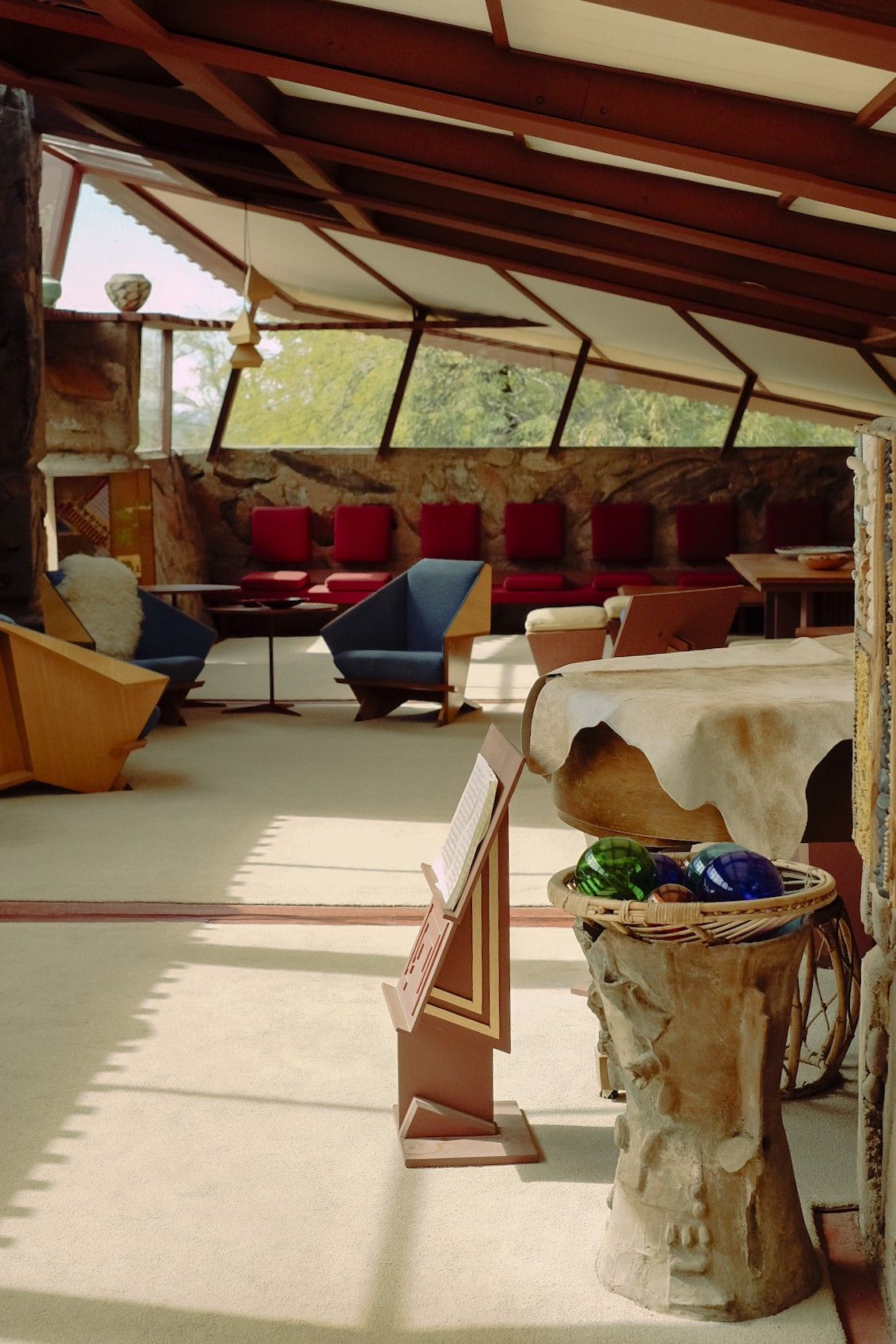A modernist home immersed in the golden greenery of a desert features wood and textile ceilings in beige, carpeted floors in the same color, and geometrical furniture in tones of blue, acid green, bordeaux, and earthy browns.