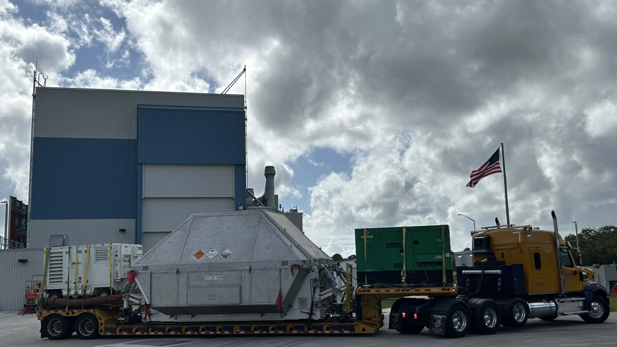 a cargo truck hauls a large flatbed trailer holding a boxy silver structure near a large building
