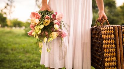 Woman wearing a white dress, holding a wicker picnic basket and a bouquet of roses