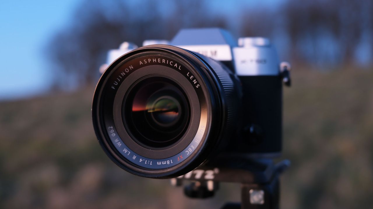 A side-on view of the Fujifilm X-M5 with the Fujifilm XF 18mm f/1.4 lens attached, with a field in the background