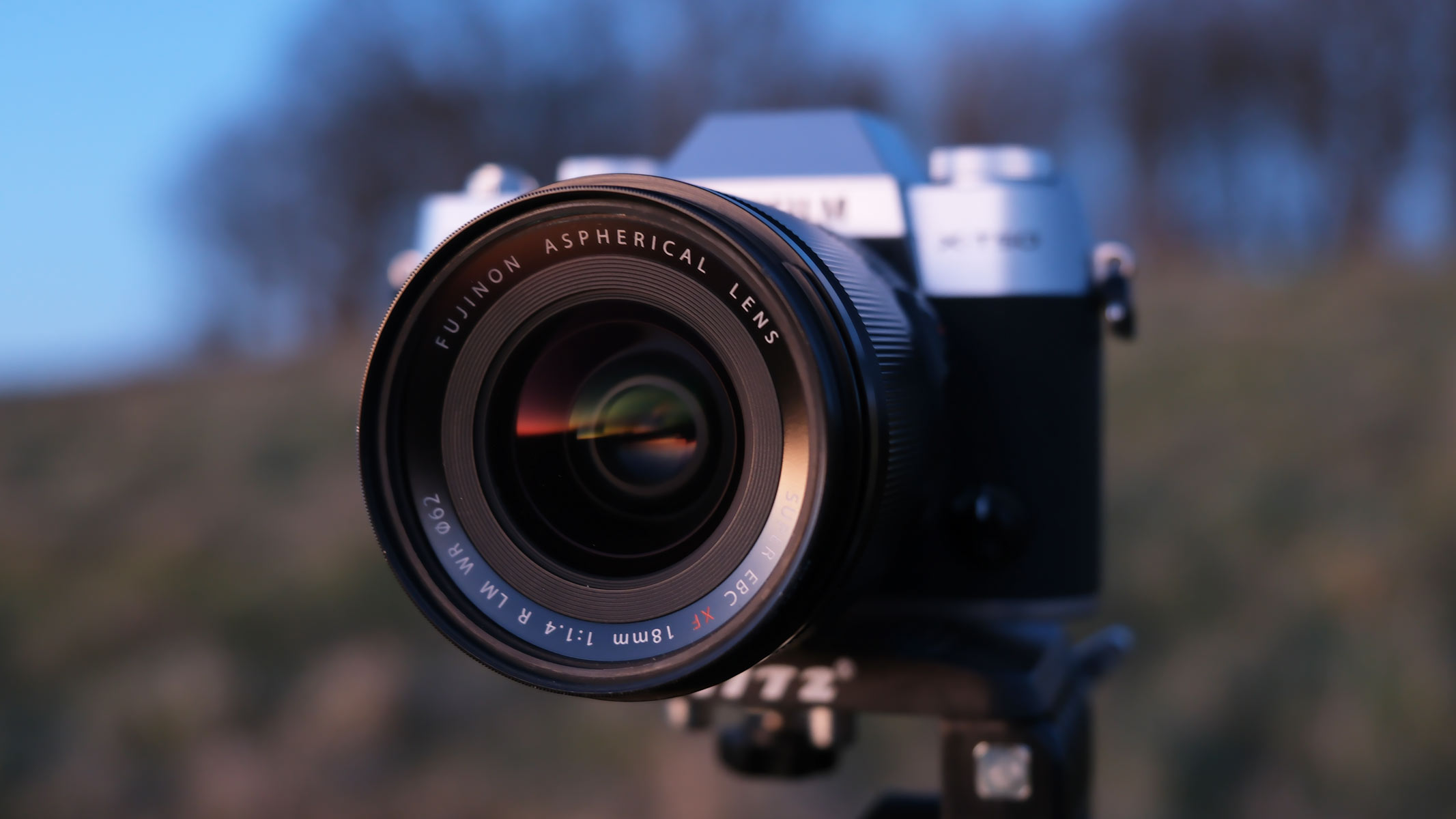 A side-on view of the Fujifilm X-M5 with the Fujifilm XF 18mm f/1.4 lens attached, with a field in the background