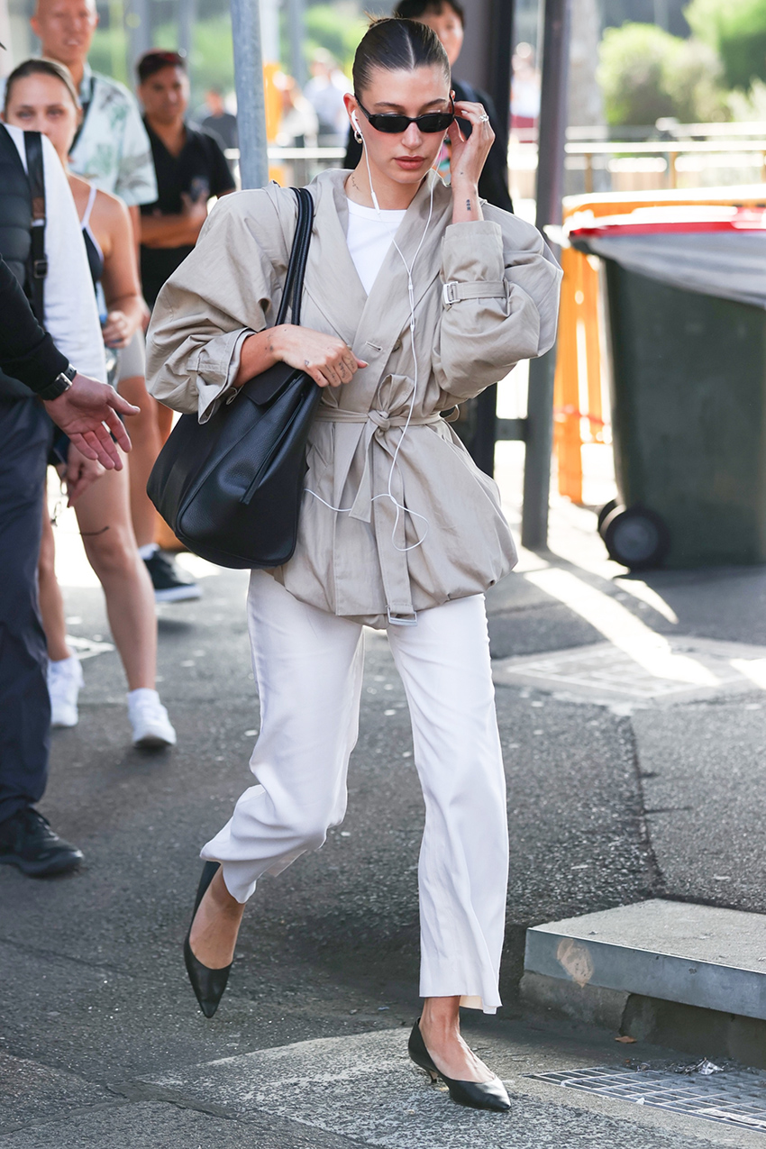 Hailey Bieber wearing a tan jacket, white pants, and kitten-heel pumps at the airport in Australia.