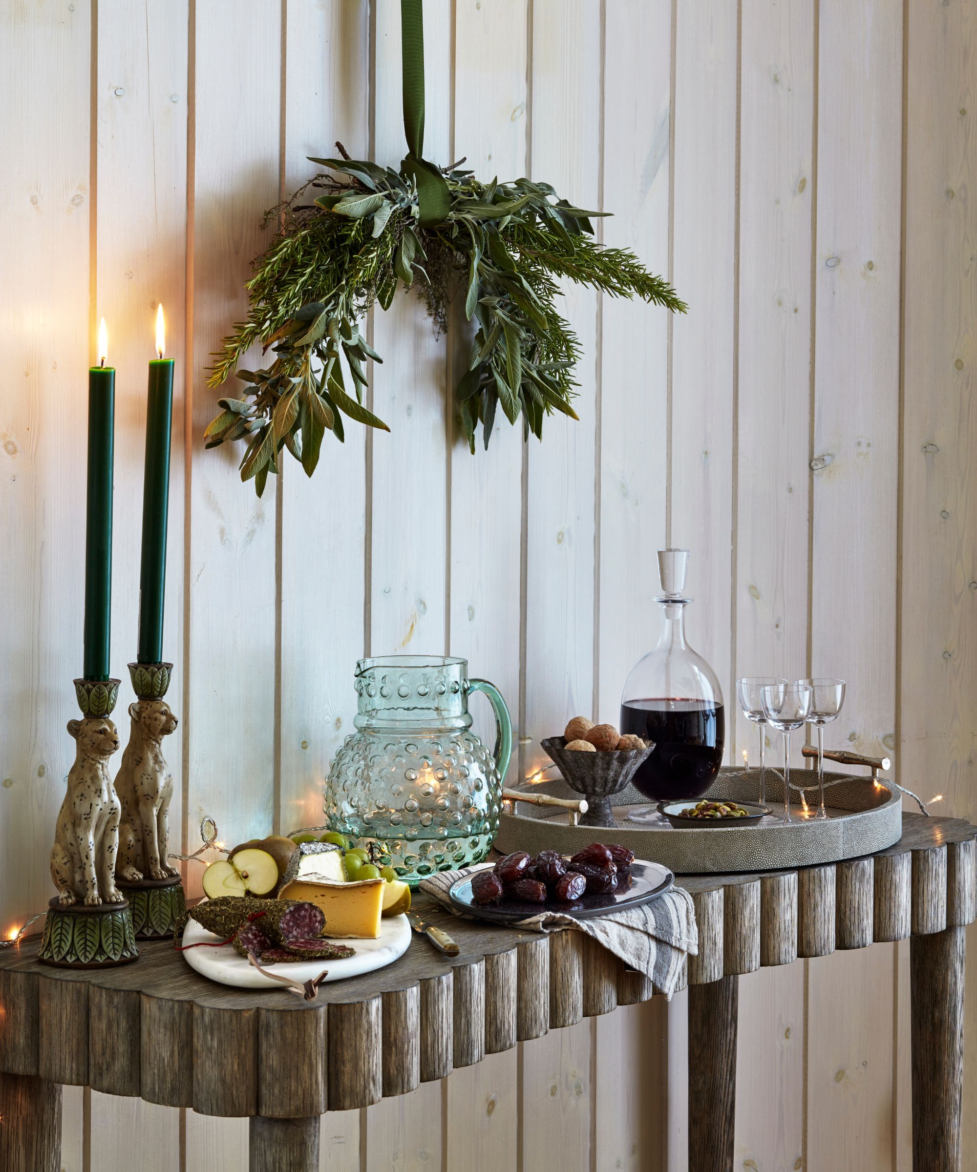 Wooden console table set with a decanter of wine and glasses, a cheese board, jug of water and taper candles with a bunch of foliage hanging above