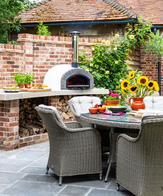 Outside dining area, with brick built pizza oven in background, woven garden furniture, vase with sunflowers,