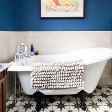 Roll top bath in a bathroom with dark blue walls and white tiles and a black and white patterned tiled floor