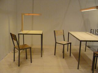 A display of chairs and table. One chair with a square shaped table featuring an attached tabled lamp AND a chair with a rectangular shaped table with a lamp
