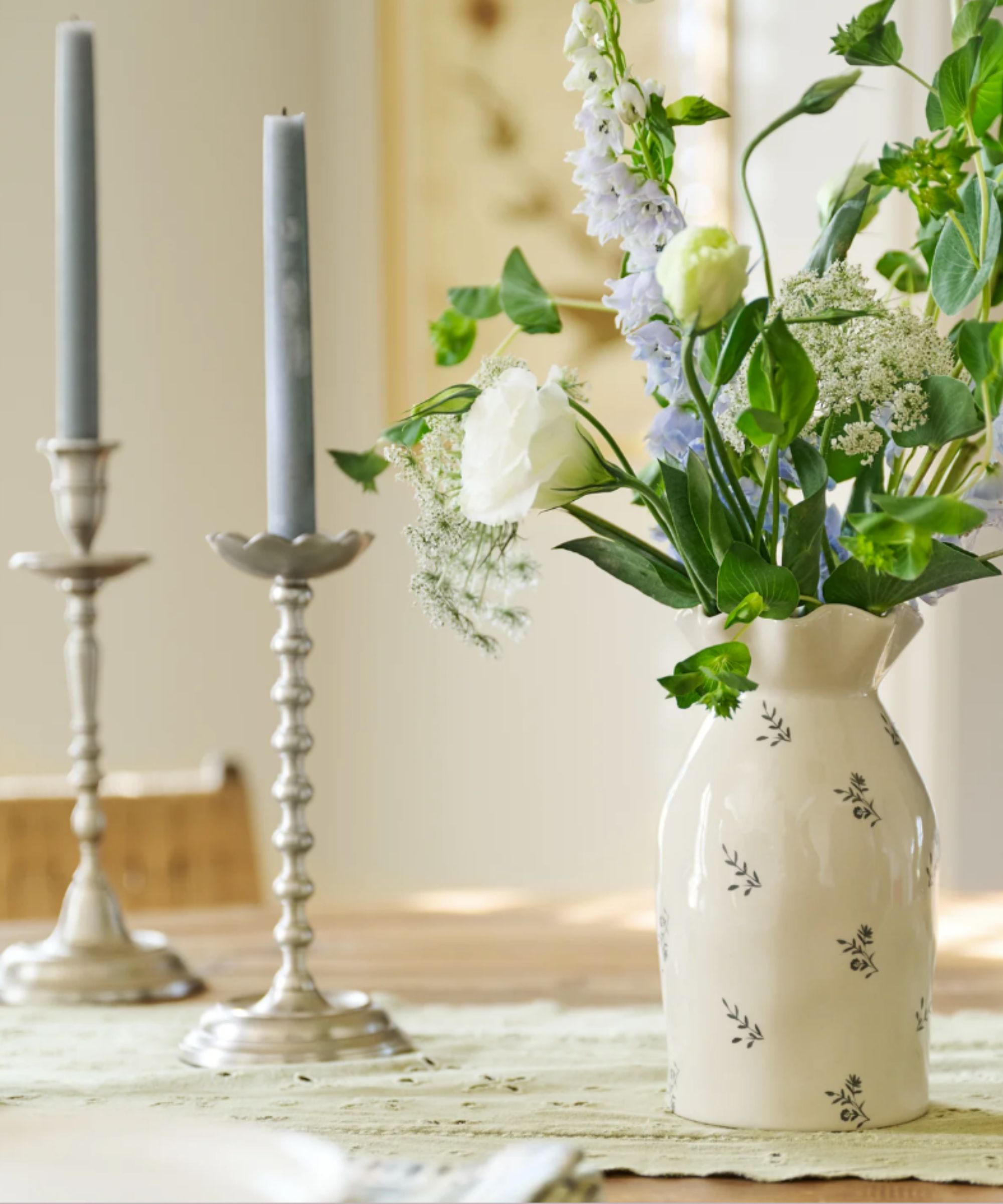 White floral ceramic vase with spring flower arrangement, next to pewter taper candle holders with pale blue tapers