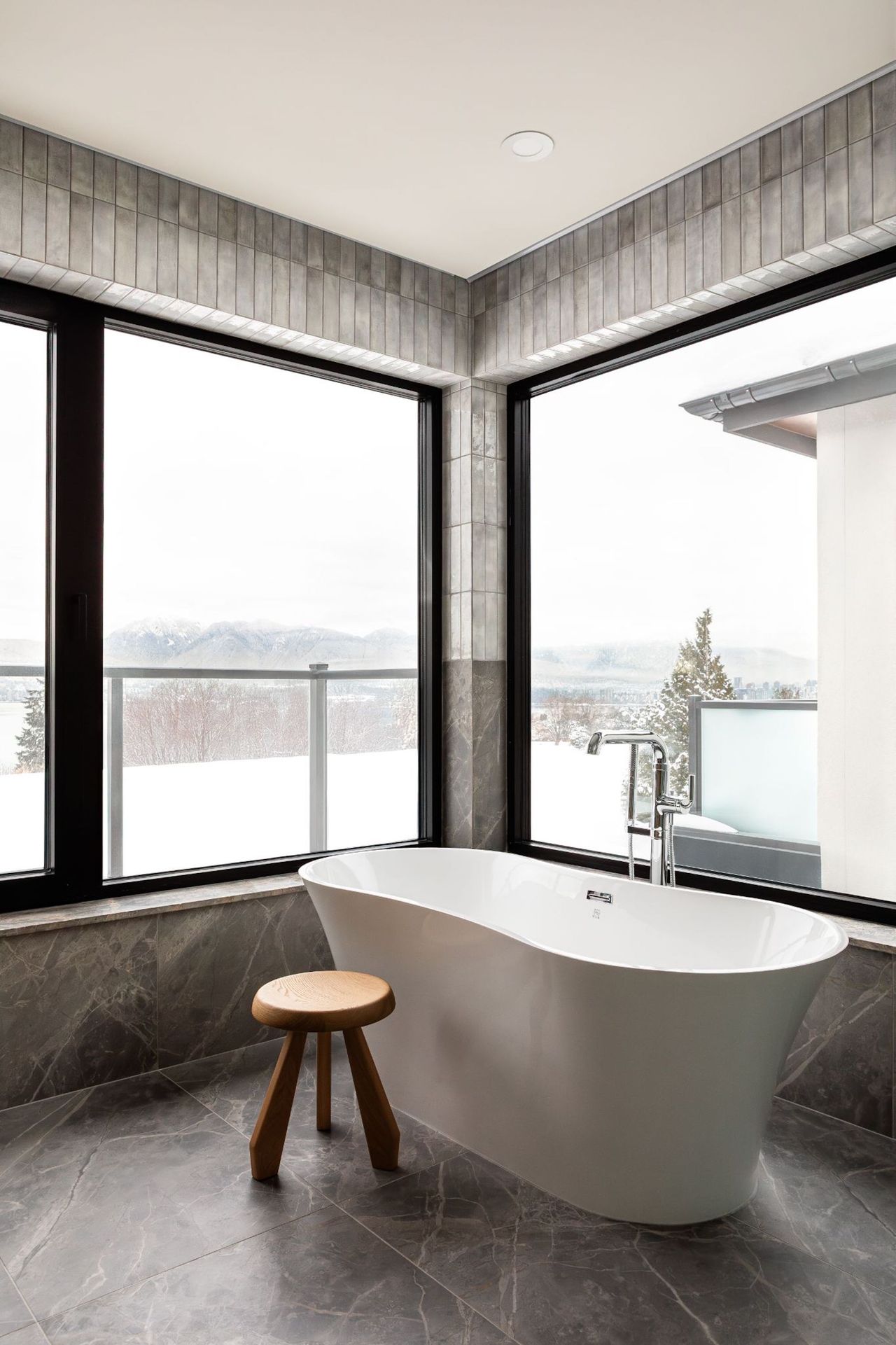 A view from the bathroom out of the large windows to the balcony and view, the interior is covered in grey tiles and marble with a free standing bathtub in front of the right window.