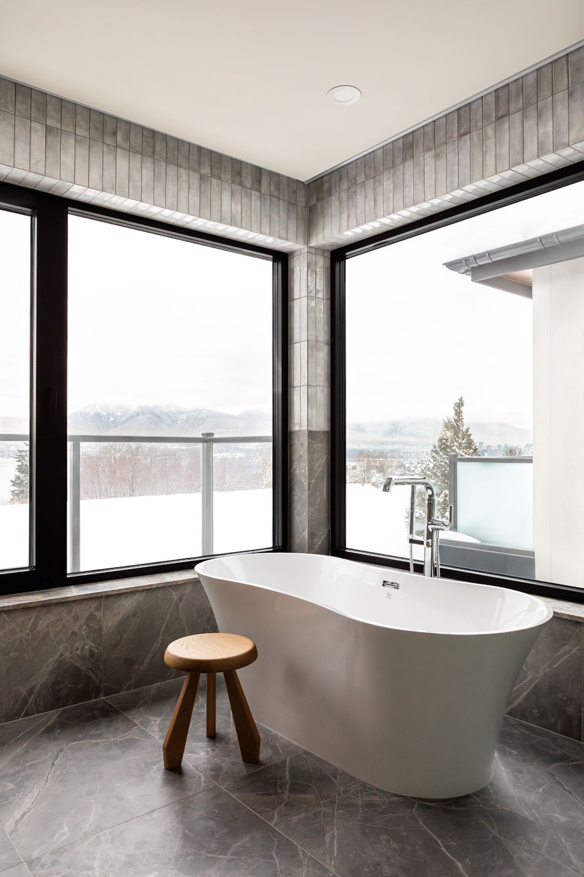 A view from the bathroom out of the large windows to the balcony and view, the interior is covered in grey tiles and marble with a free standing bathtub in front of the right window.