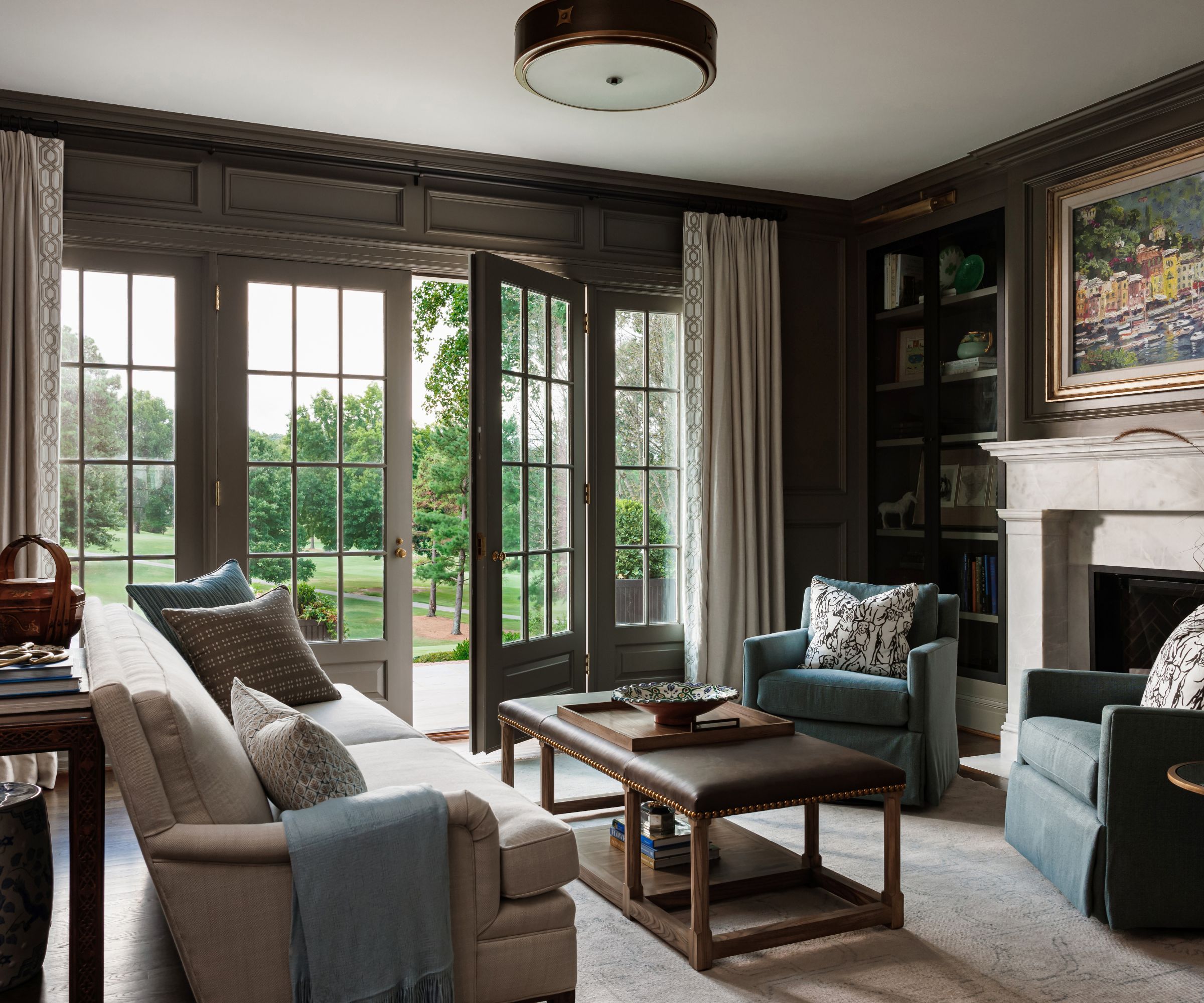 A large home office with dark gray-brown walls, French doors leading into a garden, and a cream sofa with an upholstered coffee table, and two blue armchairs in front of the fireplace.